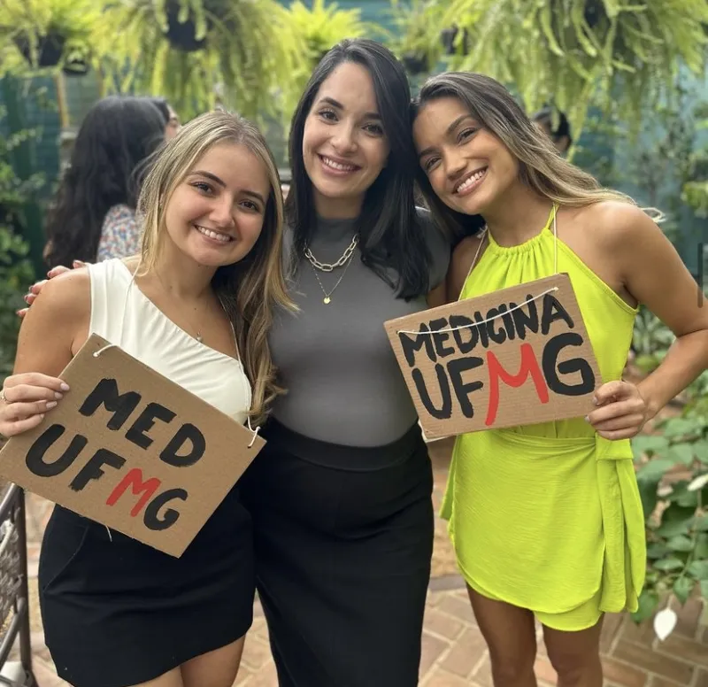 Bella Nazar entre duas alunas aprovadas em Medicina na UFMG, com placas de papelão